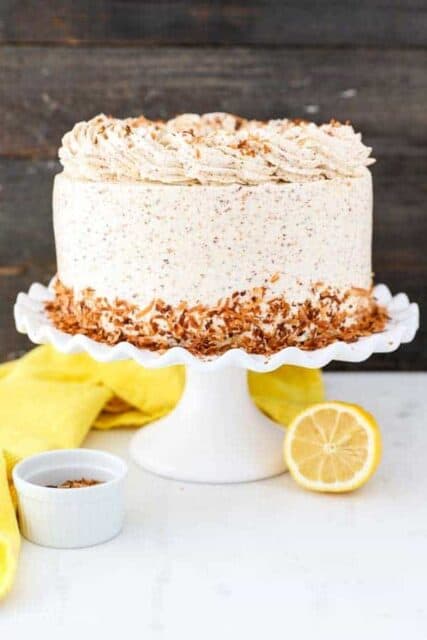 A gorgeous layer cake frosted with toasted coconut buttercream, with toasted coconut pressed into the bottom edge. It's sitting on a white ruffled cake plate, and there's a yellow napkin and sliced lemon sitting next to it.