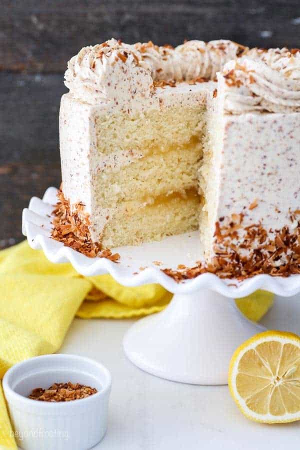 A 3 layer lemon cake with a slice missing, showing the inside of the cake which has a lemon curd filling and a toasted coconut buttercream