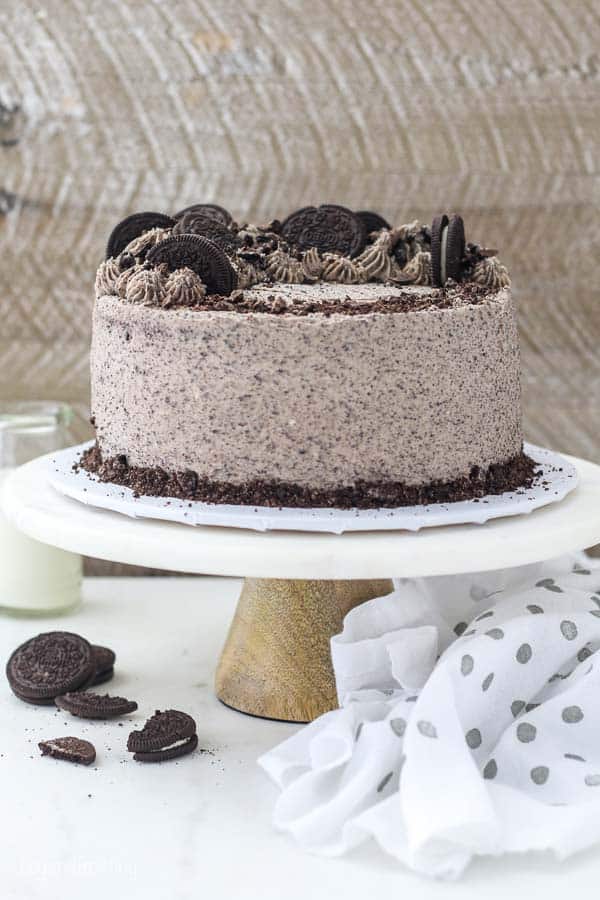 A whole Oreo cake sitting on a marble cake stand, the cake is topped with crushed Oreos. There's a white and grey polka dot cloth sitting next to the cake plate.