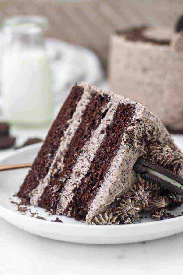 A 3 layer chocolate cake with Oreo frosting laying on a large white cake plate. The top of the slice of cake has Oreo rosettes and more cookies