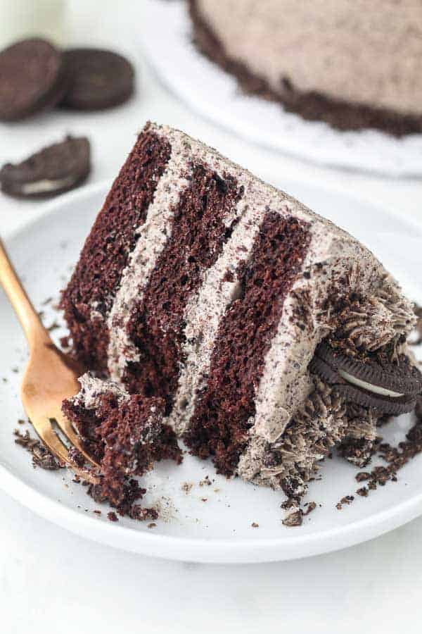 A slice of chocolate oreo cake on a white cake plate. It's 3 layers of chocolate cake with a creasy Oreo frosting.