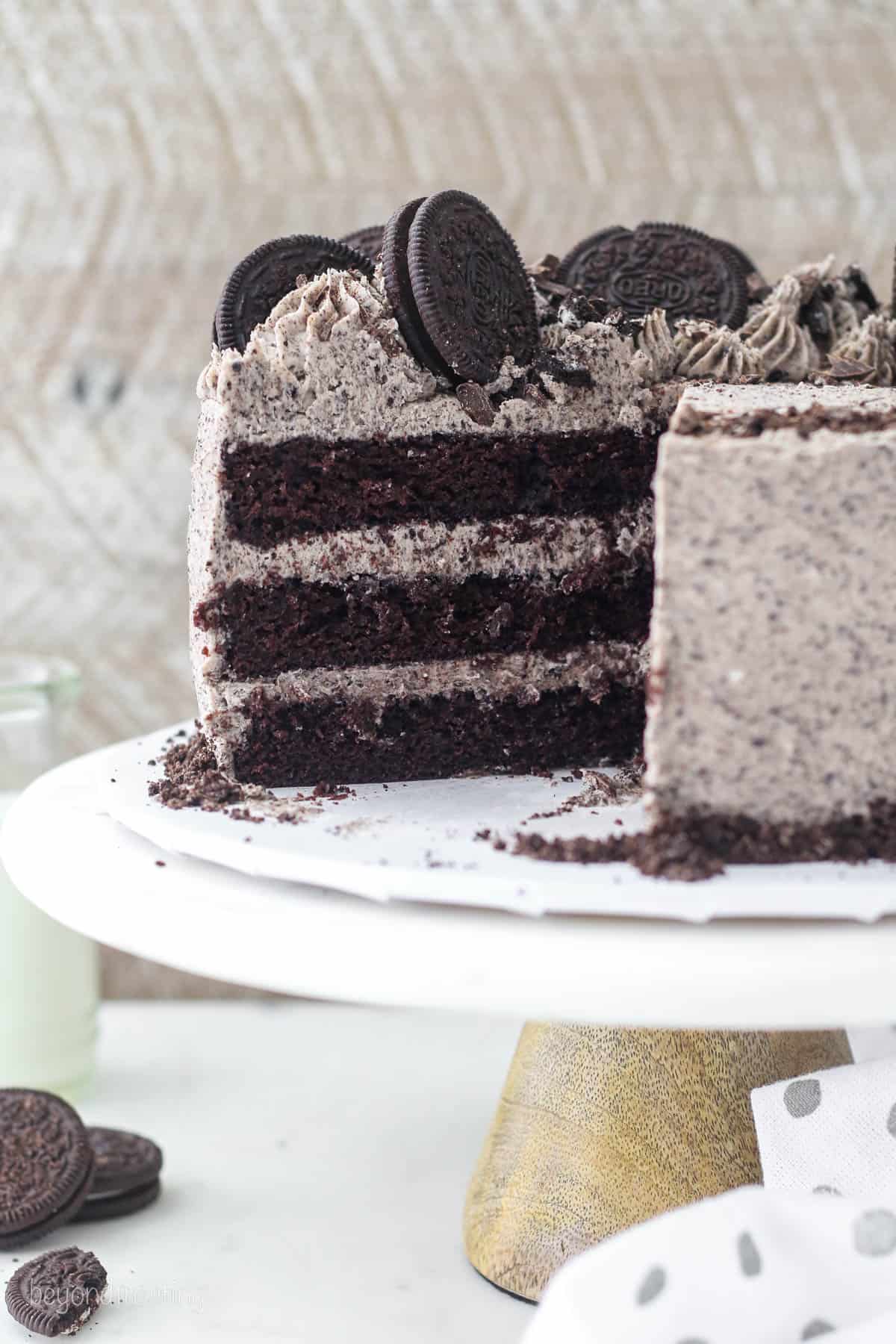 A frosted Oreo chocolate cake garnished with Oreo cookies on a cake stand, with a large slice missing.