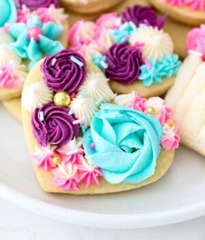 A plate of sugar cookies decorated with purple, teal, white, and pink buttercream frosting.