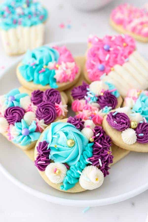 A plate full of gorgeous sugar cookies decorated with buttercream in purple, teals, white and pink frosting.