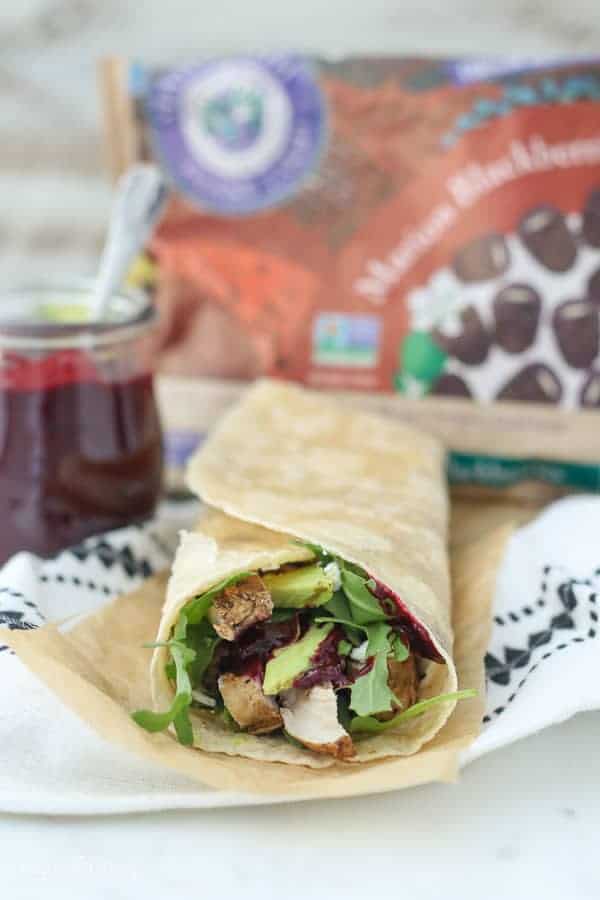 A blackberry chicken wrap is laying on a piece of brown parchment paper. Blurred out in the background is a jar of blackberry sauce and a package of frozen blackberries