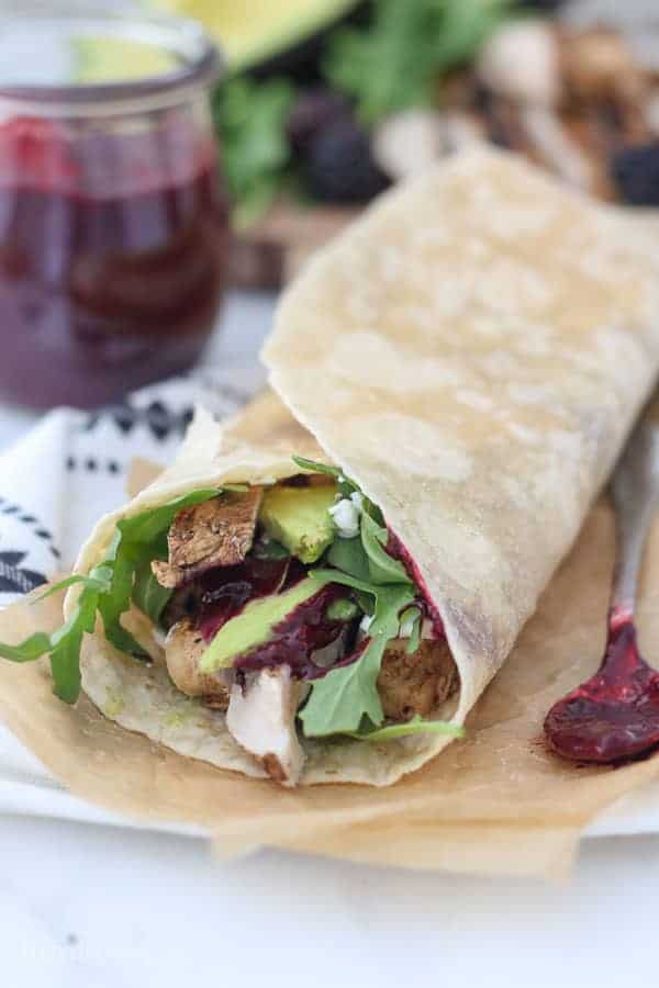 A chicken wrap is laying on a piece of brown parchment paper, hanging out of the wrap is chicken, lettuce and avocado. There's also a spoon of blackberry sauce next to it