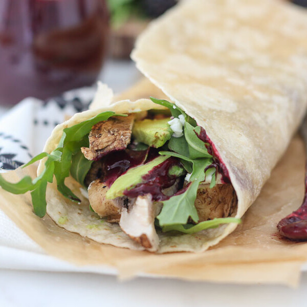 A chicken wrap is laying on a piece of brown parchment paper, hanging out of the wrap is chicken, lettuce and avocado