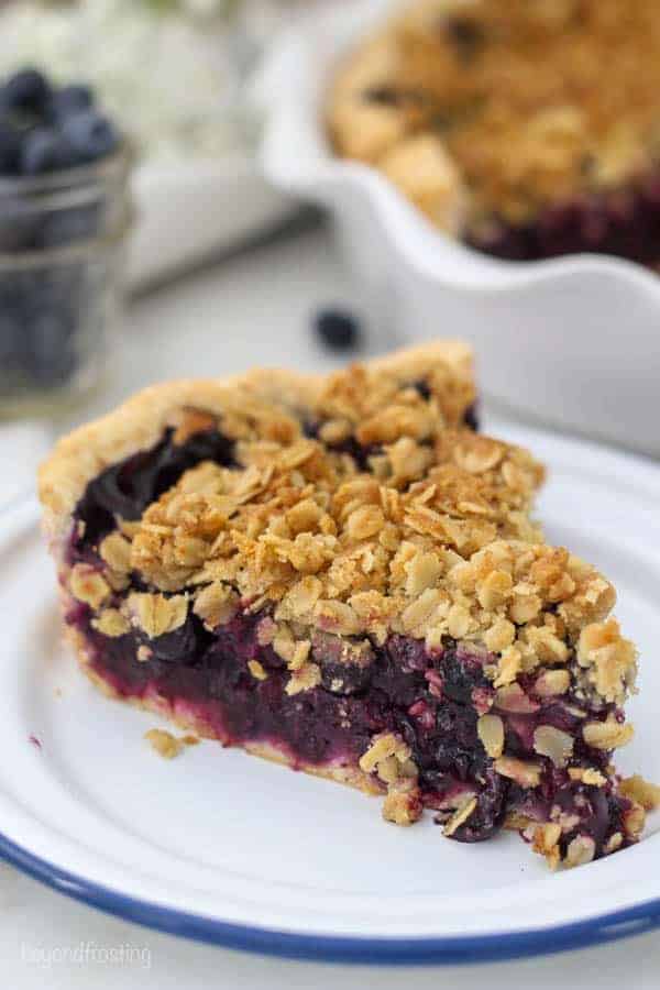 A close up shot of a slice of blueberry pie with a crumble topping sitting on a white plate with a blue rim