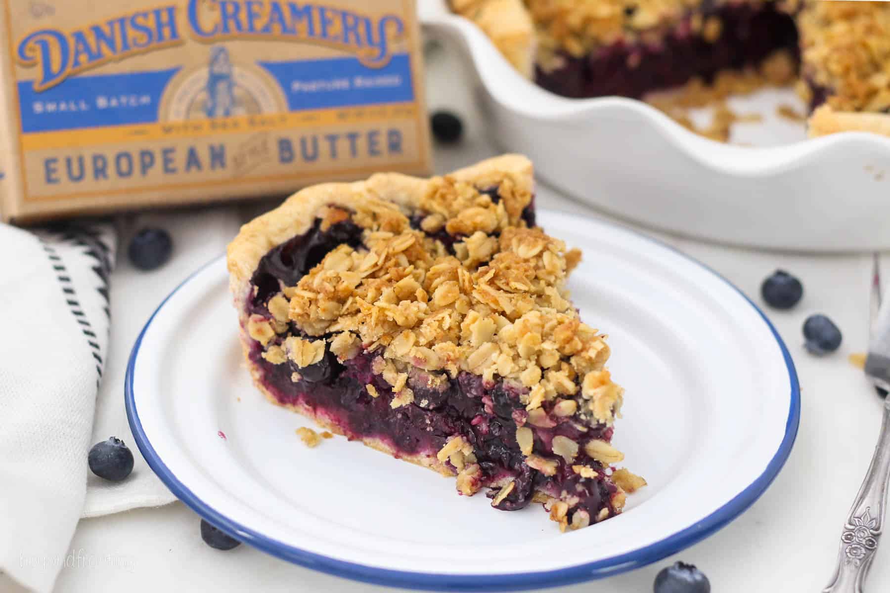 A slice of blueberry crumble pie on a white plate with a brick of butter in the background.