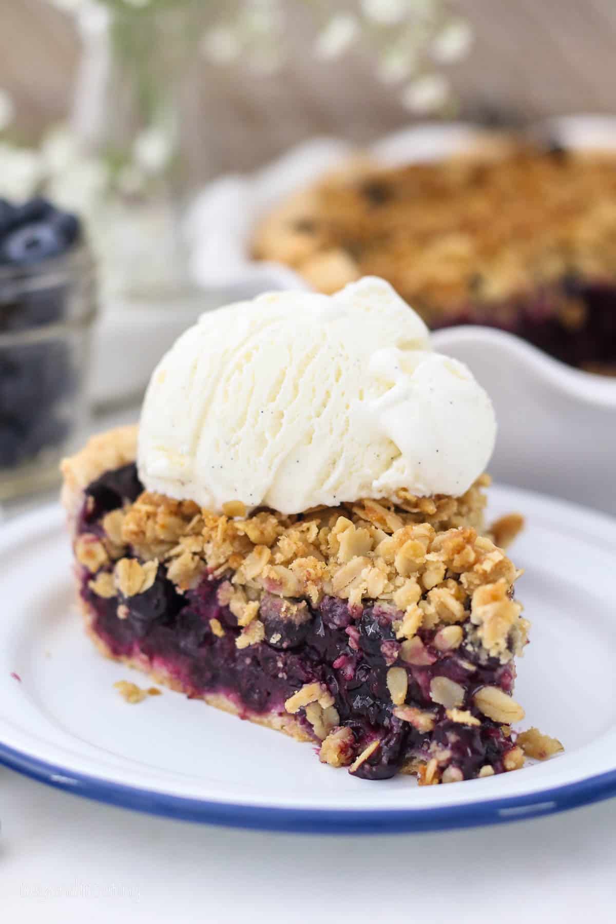 A slice of blueberry crumble pie on a white plate topped with a scoop of vanilla ice cream.