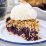 A slice of blueberry crumble pie on a white plate topped with a scoop of vanilla ice cream.