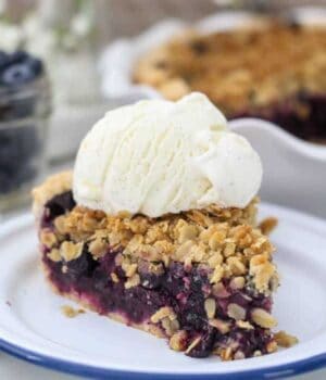 A slice of juicy blueberry pie sitting on a white plate with a blue rim, with a scoop of vanilla ice cream on top