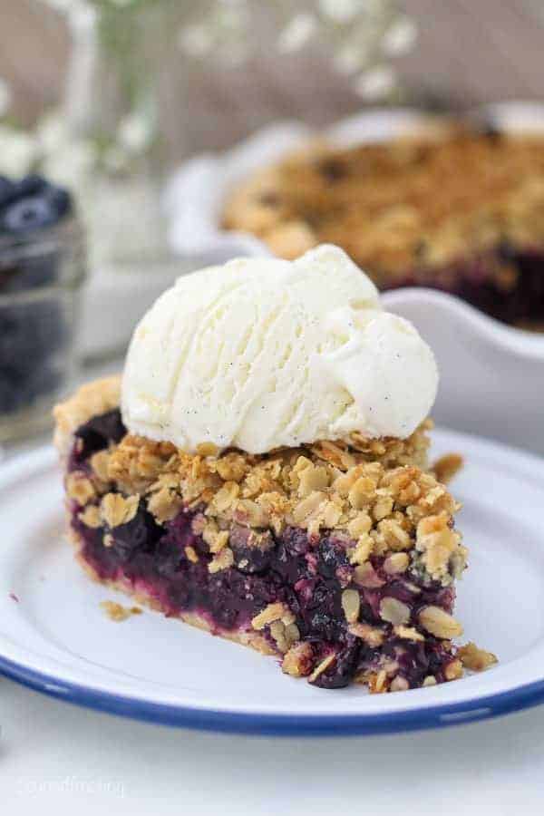 A slice of juicy blueberry pie sitting on a white plate with a blue rim, with a scoop of vanilla ice cream on top