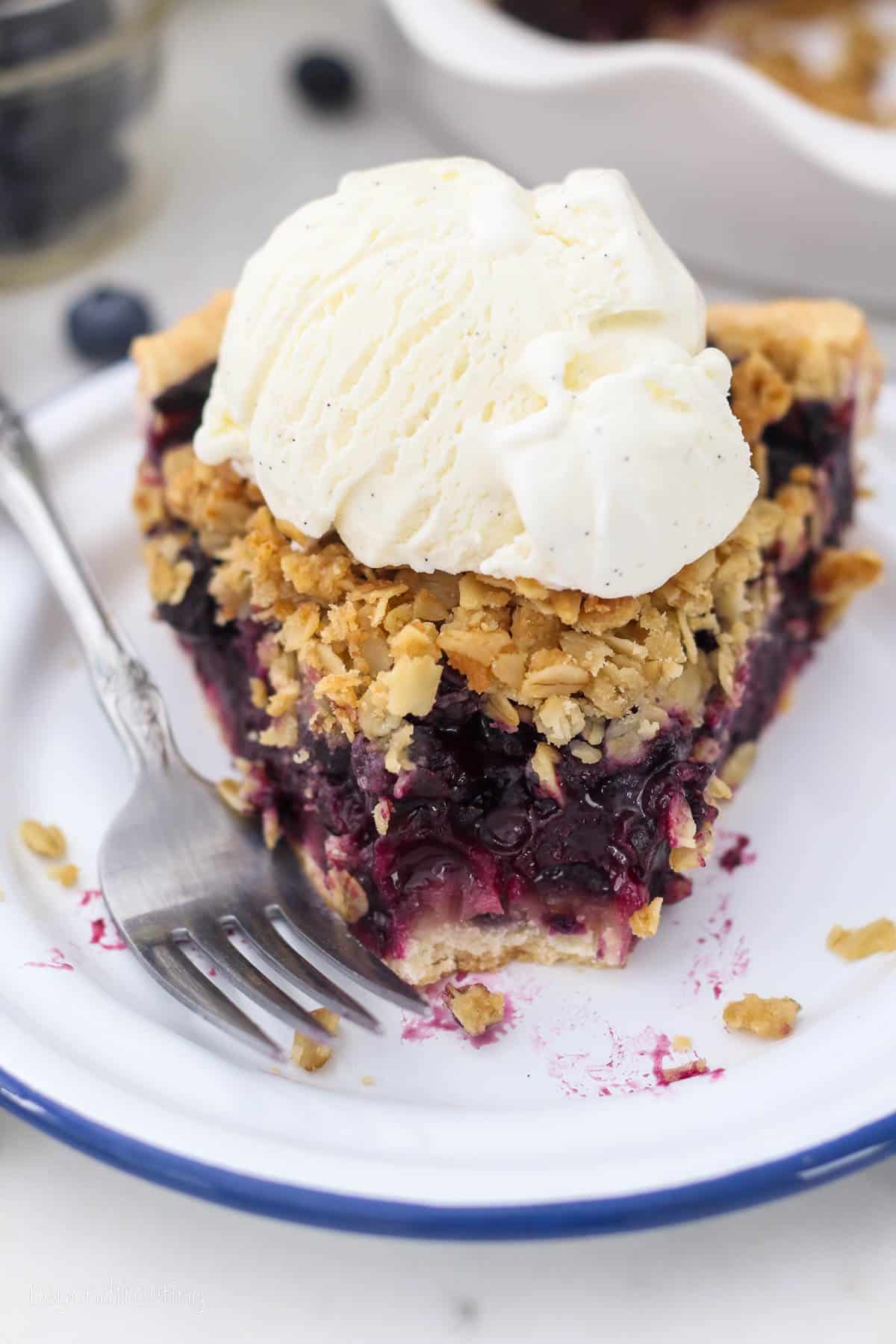 A slice of blueberry crumble pie on a white plate with a forkful missing, topped with a scoop of vanilla ice cream.