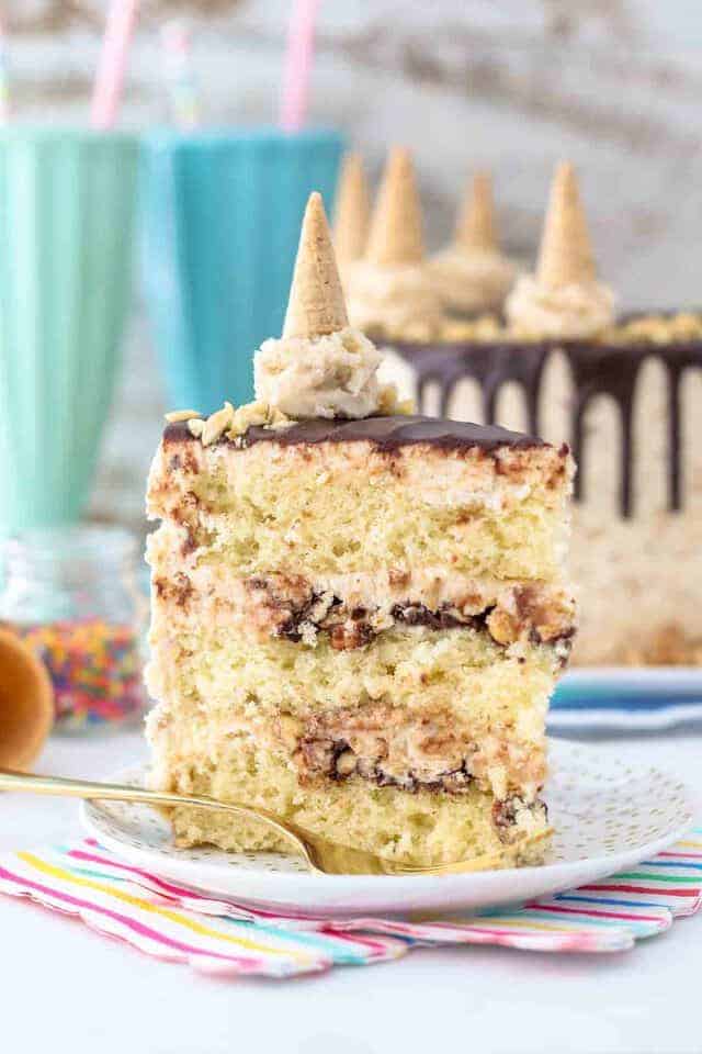 A tall slice of layered Drunmstick cake, the top of the cake has a mini ice cream cone made with frosting. You can see the layers of fudge and peanuts between the layers of cake