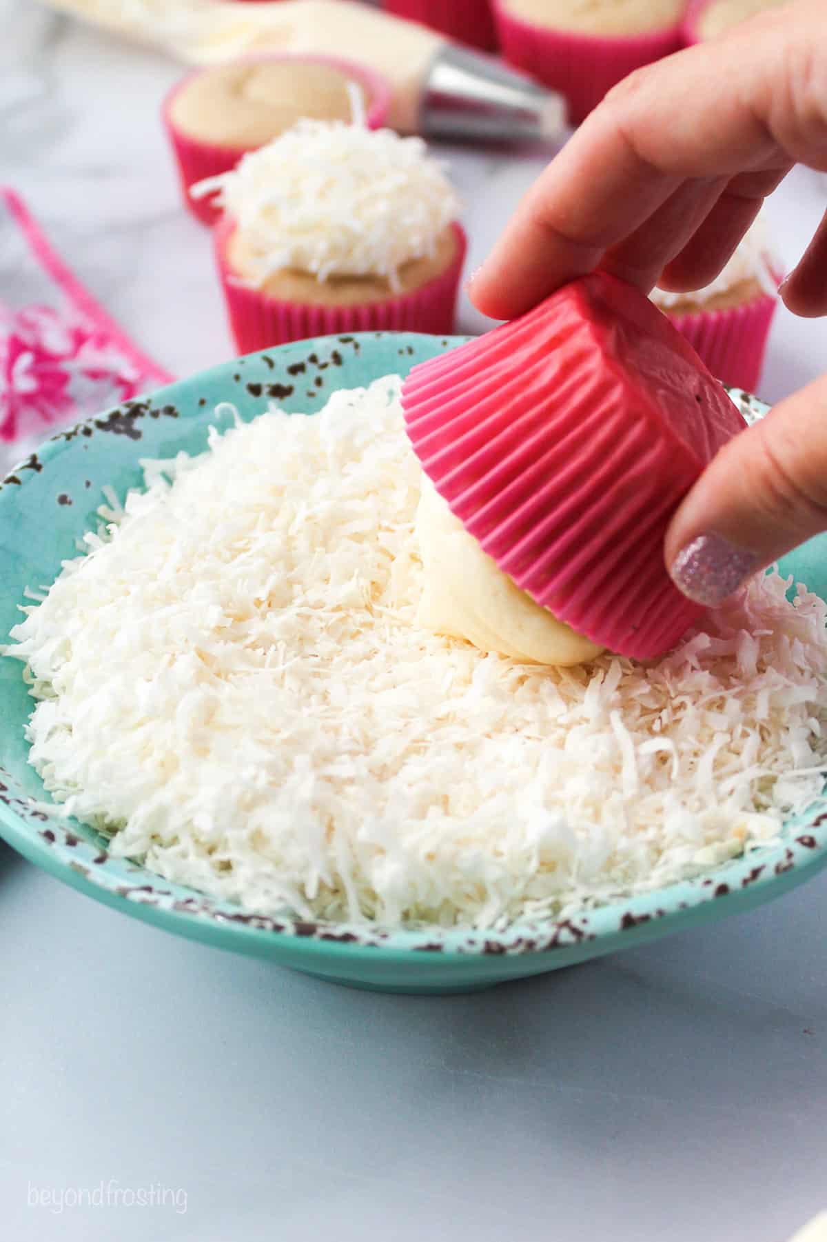 Make adorable Easter bunny cupcakes with chocolate ears following this easy step-by-step tutorial. They’re the perfect Easter cupcakes! A hand rolling the top of a frosted cupcake in a bowl of shredded coconut.