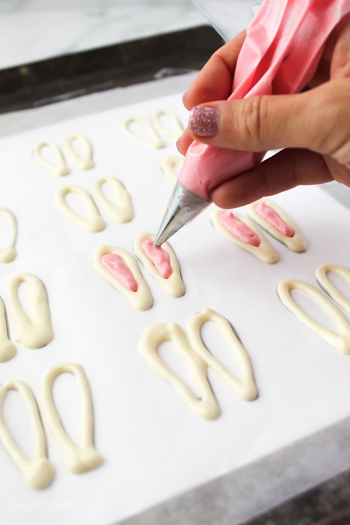 Make adorable Easter bunny cupcakes with chocolate ears following this easy step-by-step tutorial. They’re the perfect Easter cupcakes! A hand piping pink colored chocolate into the center of white chocolate shaped like bunny ears on a sheet of parchment paper.