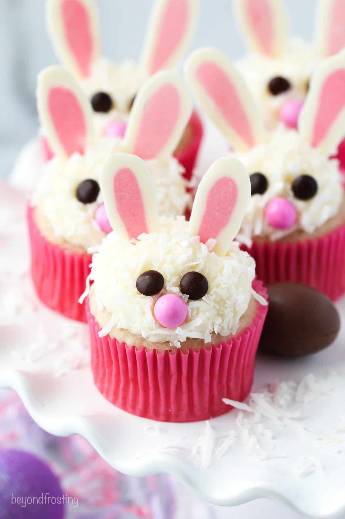 Make adorable Easter bunny cupcakes with chocolate ears following this easy step-by-step tutorial. They’re the perfect Easter cupcakes! Close up of Easter bunny cupcakes in pink liners on a cake stand.