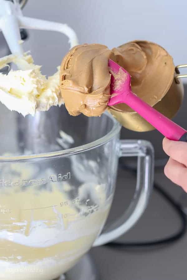 A pink spatula is spooning a big dollop of peanut butter into a mixing bowl