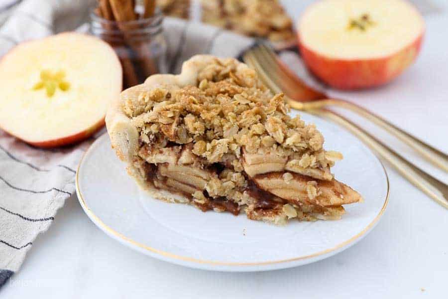 A white plate with a gold rim has a slice of apple pie with a crumble topping.