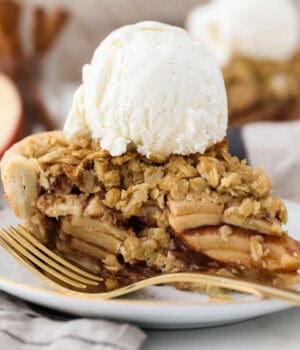 A slice of apple crumble pie topped with a scoop of vanilla ice cream on a plate, next to a fork.