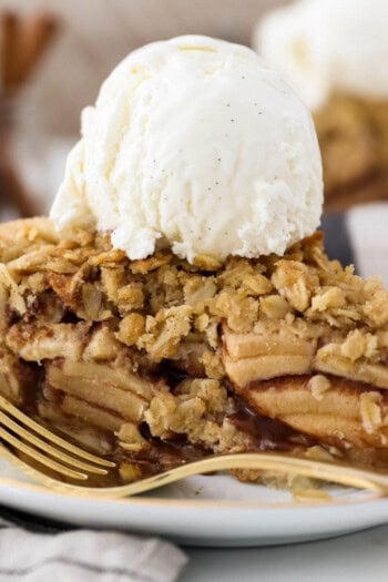 A slice of apple crumble pie topped with a scoop of vanilla ice cream on a plate, next to a fork.