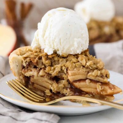 A slice of apple crumble pie topped with a scoop of vanilla ice cream on a plate, next to a fork.