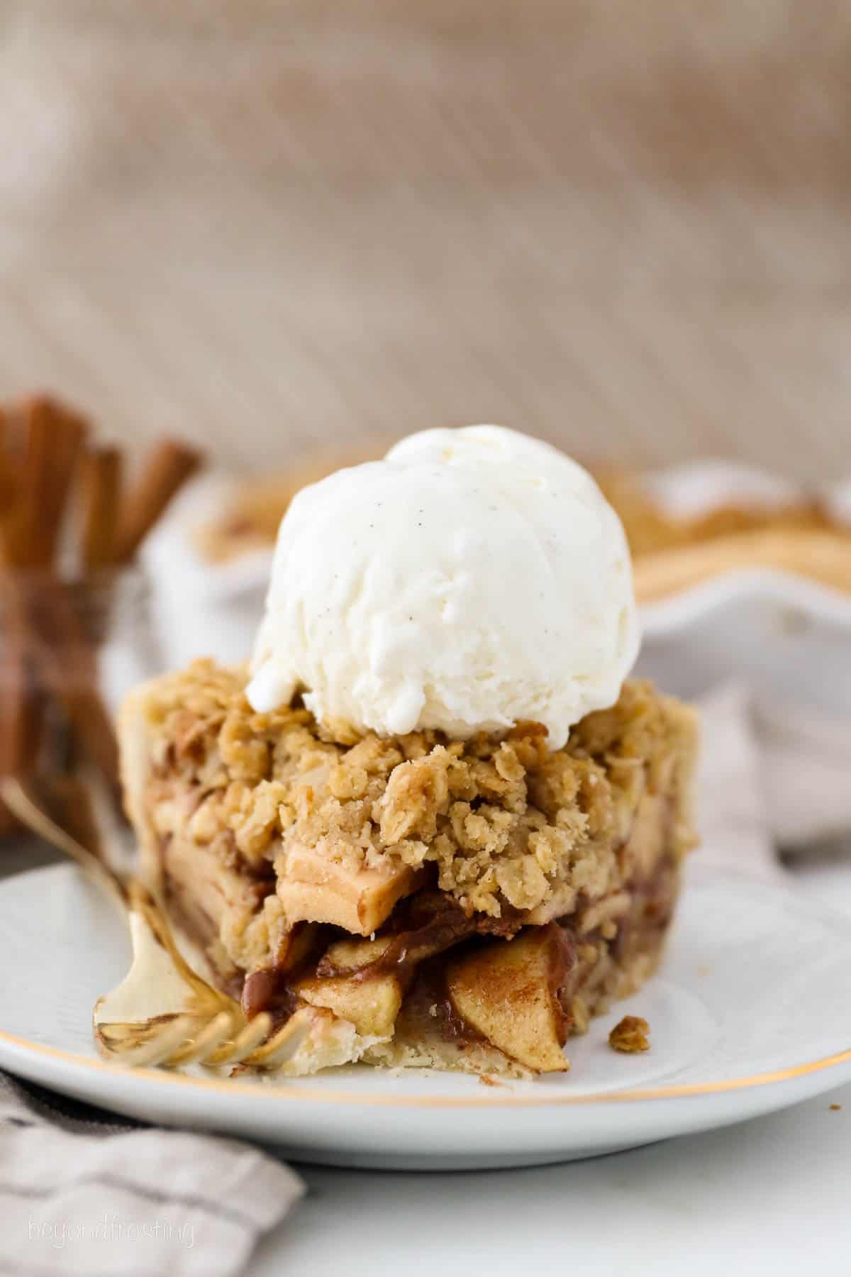 A slice of apple crumble pie on a plate with a forkful missing from the tip, topped with a scoop of vanilla ice cream.