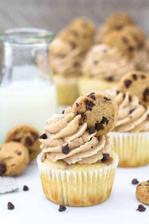 A bunch of pretty cupcake with a beautiful swirl of frosting and mini chocolate chip cookie on top. The cupcakes are surrounded with mini chocolate chips and more cookies.