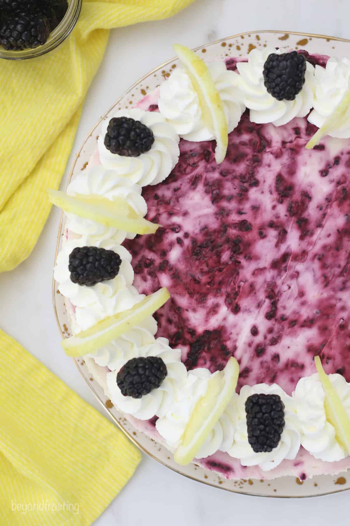 Overhead view of a blackberry lemon cheesecake decorated with swirls of whipped cream and garnished with lemon wedges and fresh blackberries.