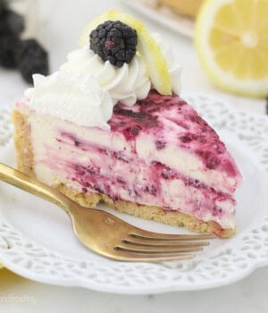 A slice of lemon blackberry cheesecake topped with whipped cream swirls and a fresh blackberry on a plate next to a fork.