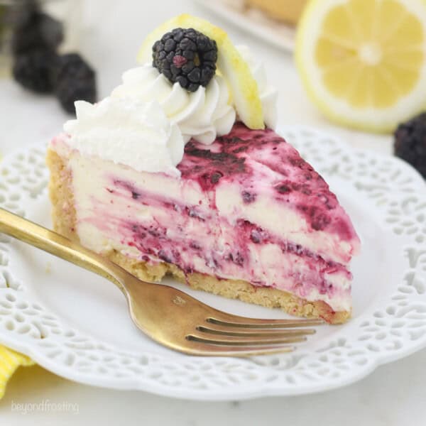 A slice of lemon blackberry cheesecake topped with whipped cream swirls and a fresh blackberry on a plate next to a fork.