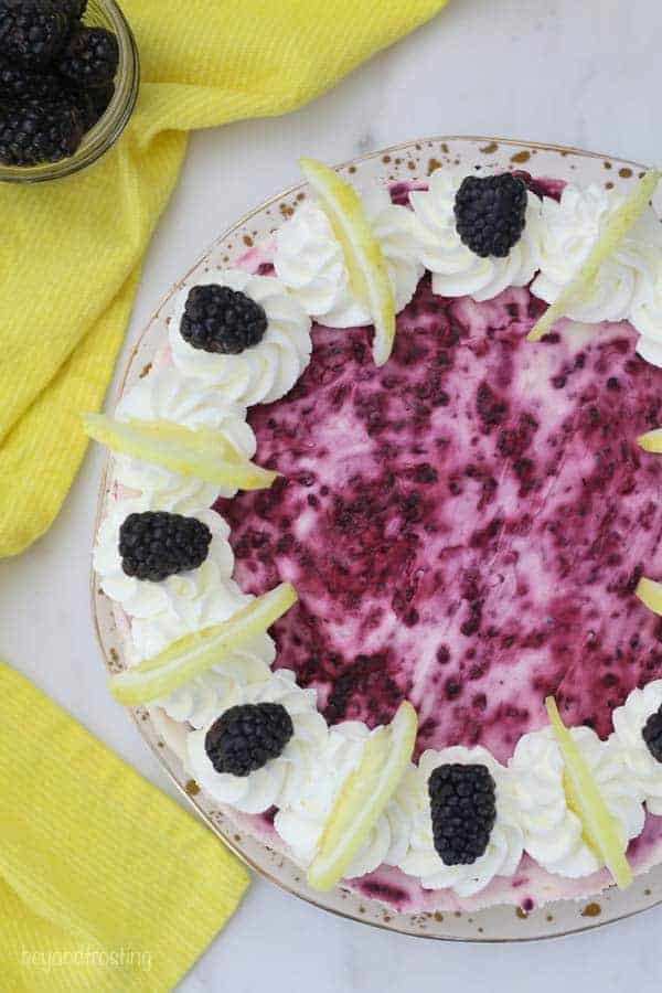 A top down view of a No-Bake Blackberry Lemon Cheesecake showing the gorgeous purple color on top from the blackberries. The cheesecake is garnished with sliced lemons and blackberries with swirls of whipped cream.