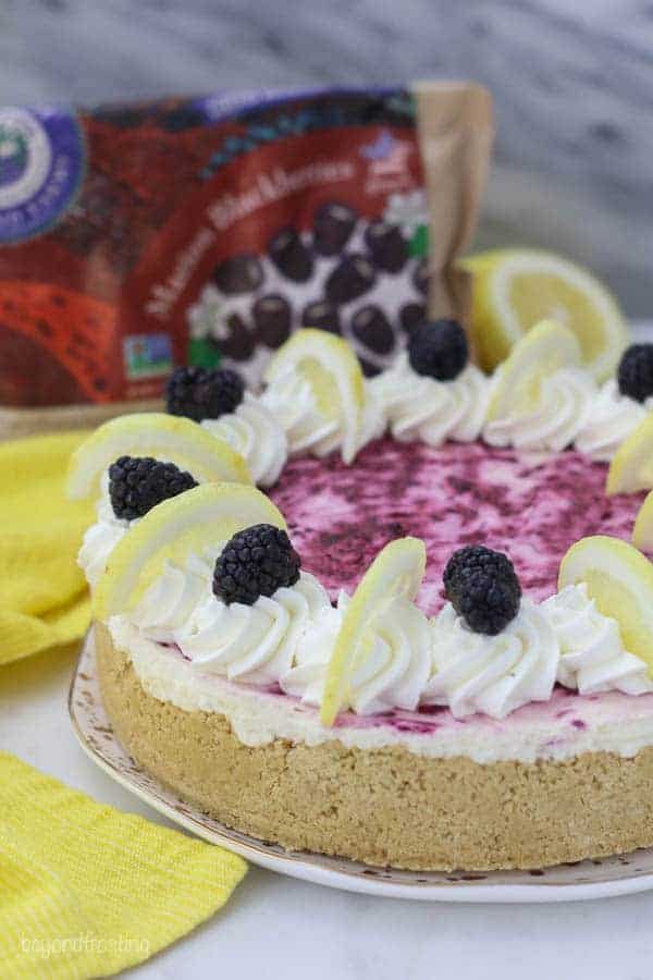 A no-bake lemon cheesecake with a beautiful swirl of blackberry on top is garnished with whipped cream, sliced lemons and fresh blackberries on top. There's a bag of frozen blackberries blurred out in the background