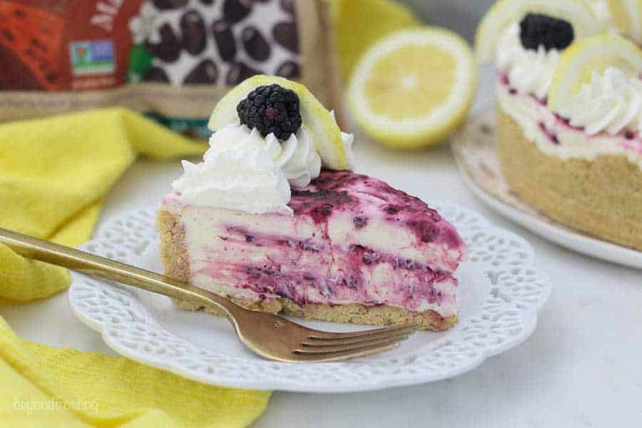 A no-bake lemon cheesecake with swirls of blackberries, it's got a bite taken out of it, and a gold fork sitting on a white plate. There's a bag of frozen Oregon blackberries blurred out in the background