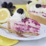 A no-bake lemon cheesecake with swirls of blackberries, it's got a bite taken out of it, and a gold fork sitting on a white plate. Blurred out in the background in the rest of the cheesecake, a lemon and some blackberries.