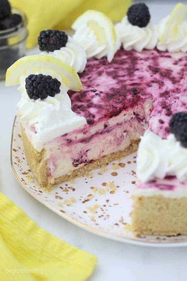 A whole no-bake lemon cheesecake with a slice missing showing the inside of the cheesecake which has swirls of blackberry sauce. You can also see the top of the cheesecake has a gorgeous purple color from the berries