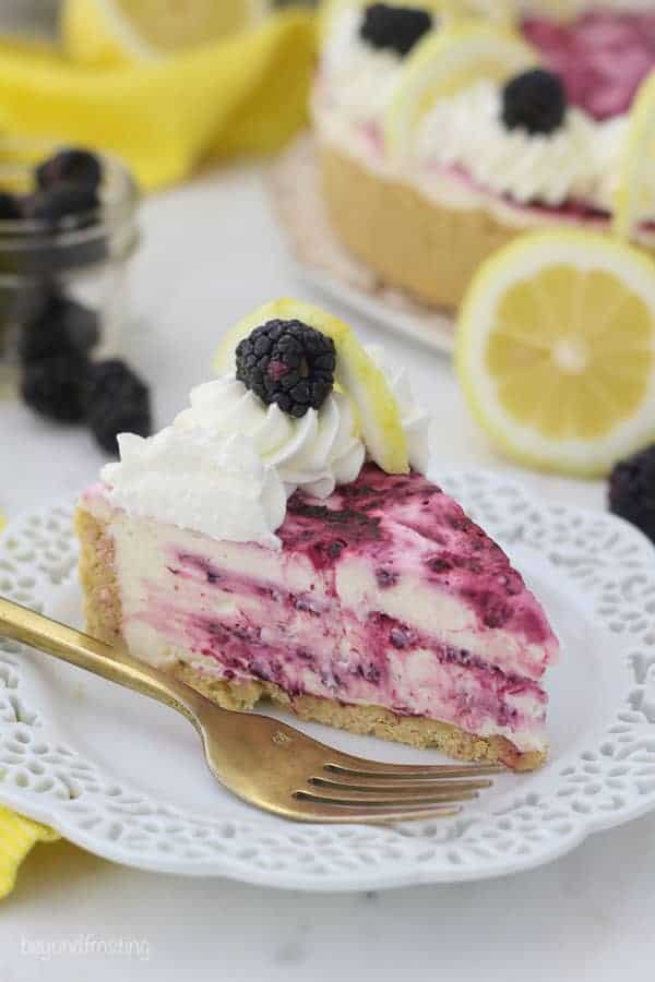 A gorgeous 3/4 view of a slice of blackberry swirled lemon cheesecake on a white plate with a gold fork sitting next to the slice of cheesecake. There's lemons and blackberries blurred out in the background