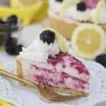 A slice of cheesecake with a blackberry swirl sitting on white plate with a ruffled edge and a gold fork. There's lemons and blackberries blurred out in the background