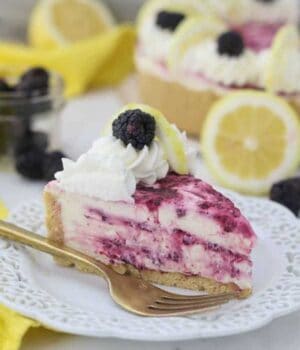 A slice of cheesecake with a blackberry swirl sitting on white plate with a ruffled edge and a gold fork. There's lemons and blackberries blurred out in the background