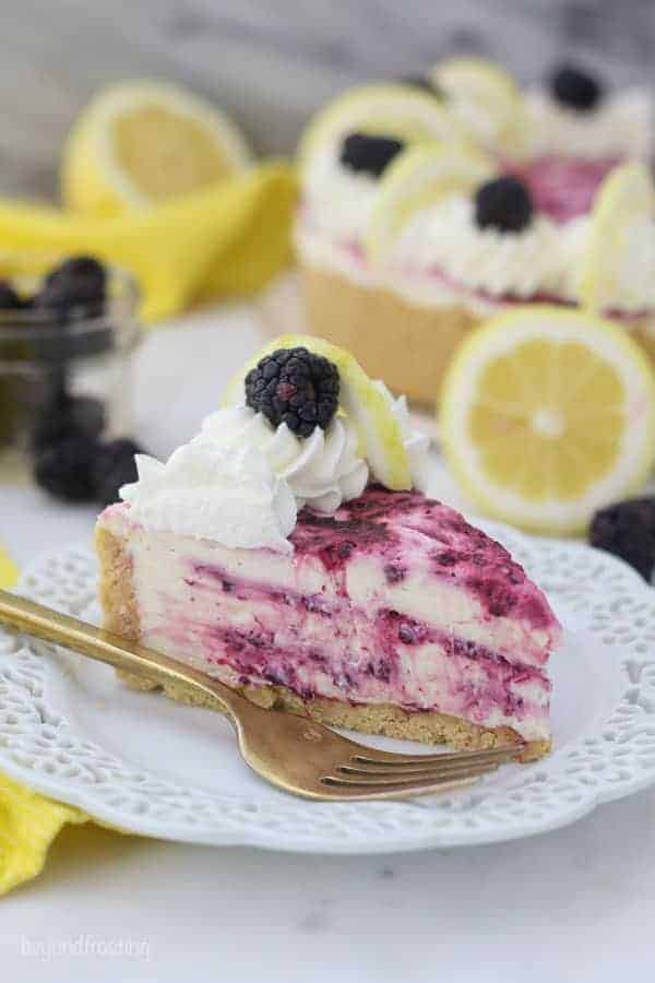 A slice of cheesecake with a blackberry swirl sitting on white plate with a ruffled edge and a gold fork. There's lemons and blackberries blurred out in the background