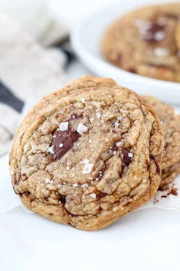 A chocolate chip cookie leaning up against a smaller stack of cookies, with giant chunks of chocolate and sea salt on top