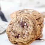 A Cookie is leaning up against a stack of other cookies. There's a big chocolate chunk on top and flaky sea salt