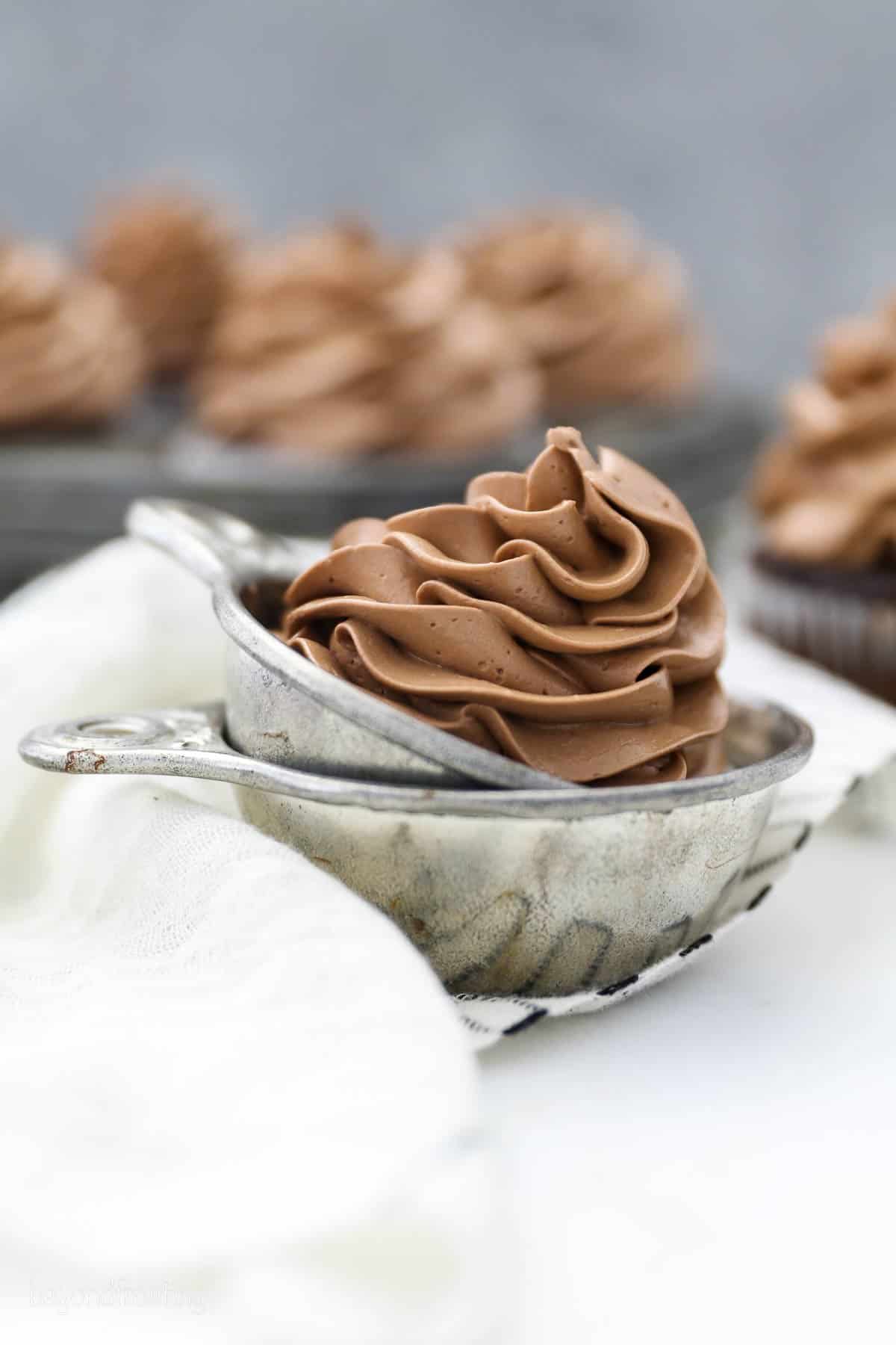 Chocolate Swiss meringue buttercream swirled in a silver serving dish with a pan of cupcakes in background.