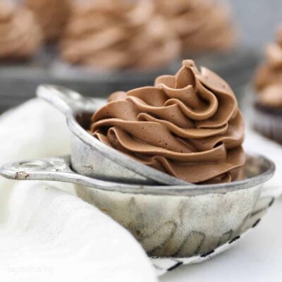 Chocolate Swiss meringue buttercream swirled in a silver serving dish with pan of frosted cupcakes out of focus.
