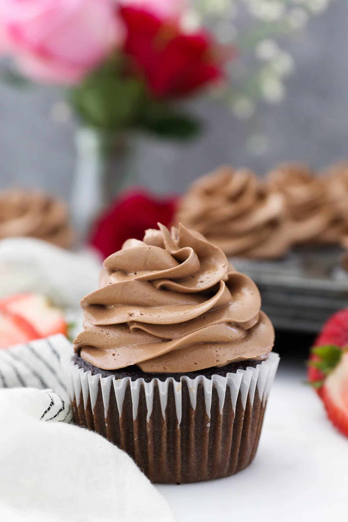 One chocolate cupcake frosted with chocolate Swiss meringue buttercream with a pan of more cupcakes in background.