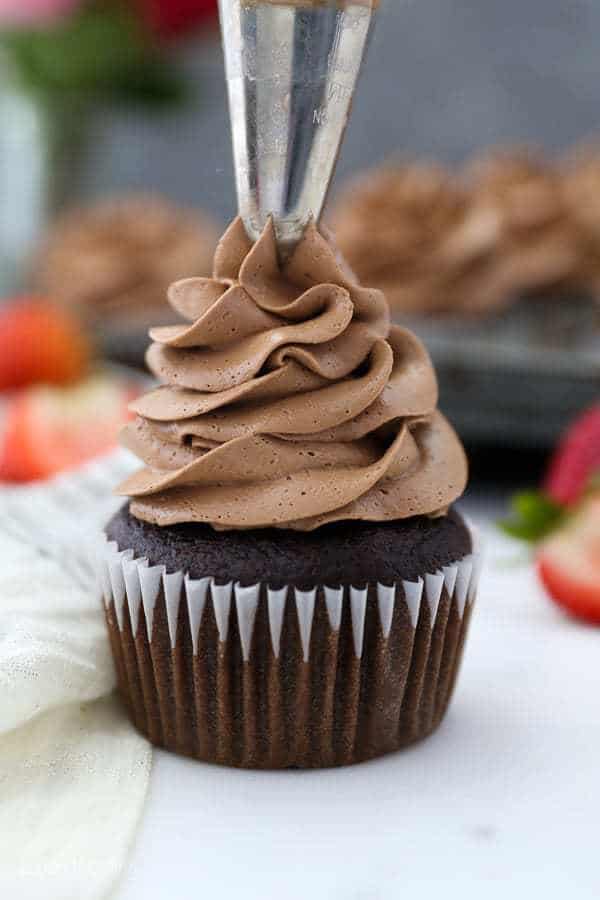 A piping bag filled with Chocolate Swiss Meringue Buttercream is piping frosting onto a chocolate cupcake