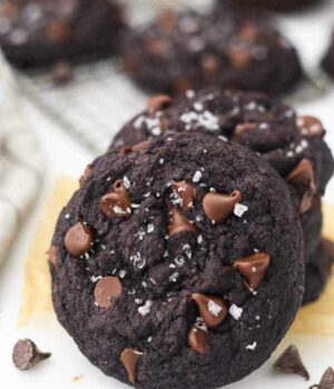 A chocolate cookie leaning up against a stack of more cookies. The top of the cookie is dotted with chocolate chips and sea salt