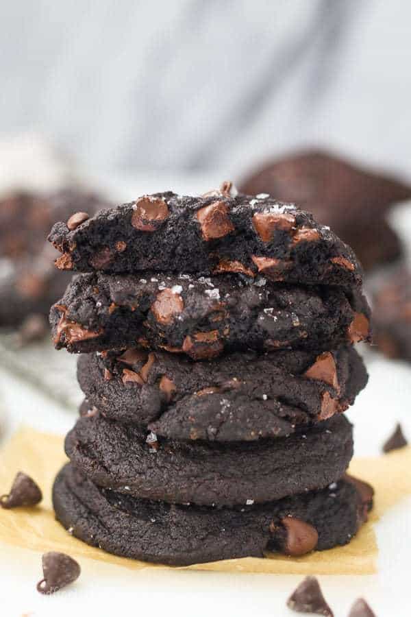 Stacks of dark chocolate cookies with chocolate chips. The top two cookies are broken in half showing the inside
