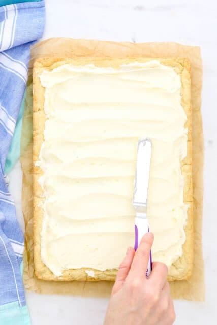 A hand uses an offset spatula to spread frosting over a slab of uncut sugar cookie bars.